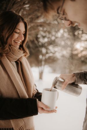 Person in beige sweater smiling while holding white mug outdoors with soft bokeh lighting in background.