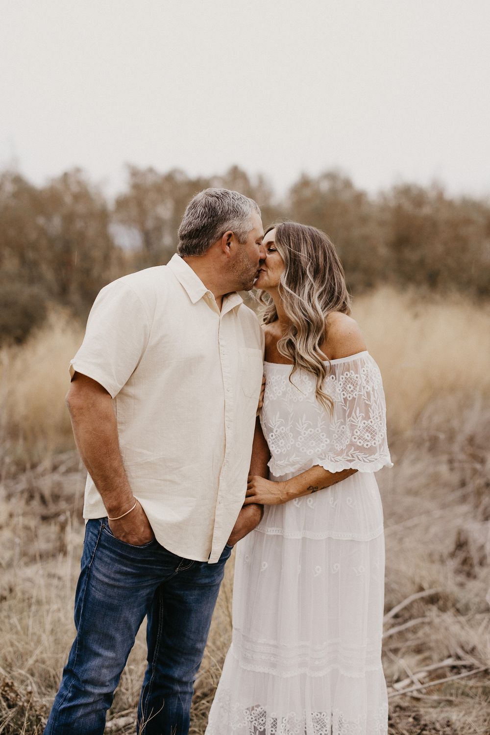 A romantic couple in white outfits share tender moments in a rustic field setting at sunset.