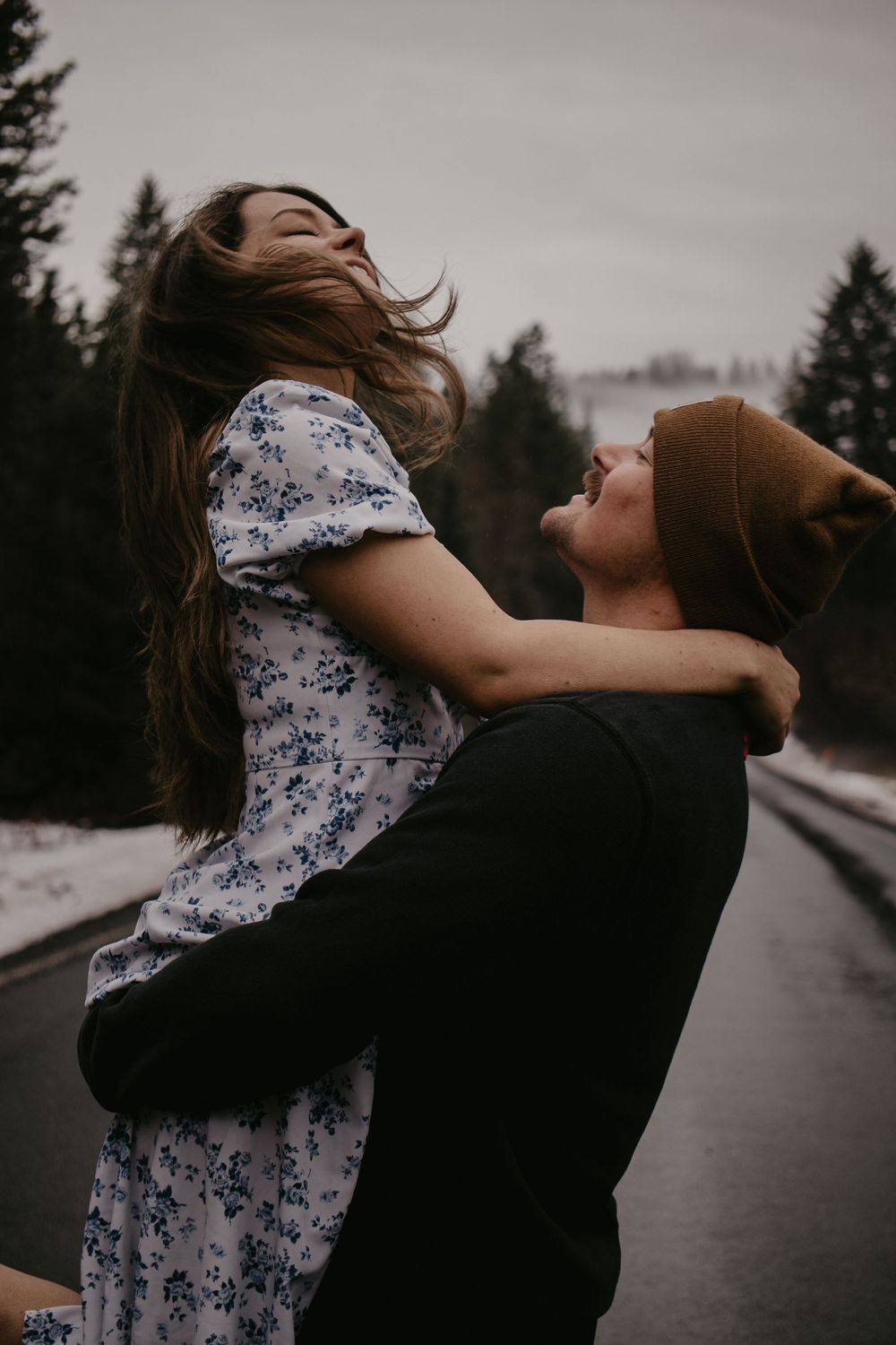 Couple embracing on a rainy road with pine trees in background.