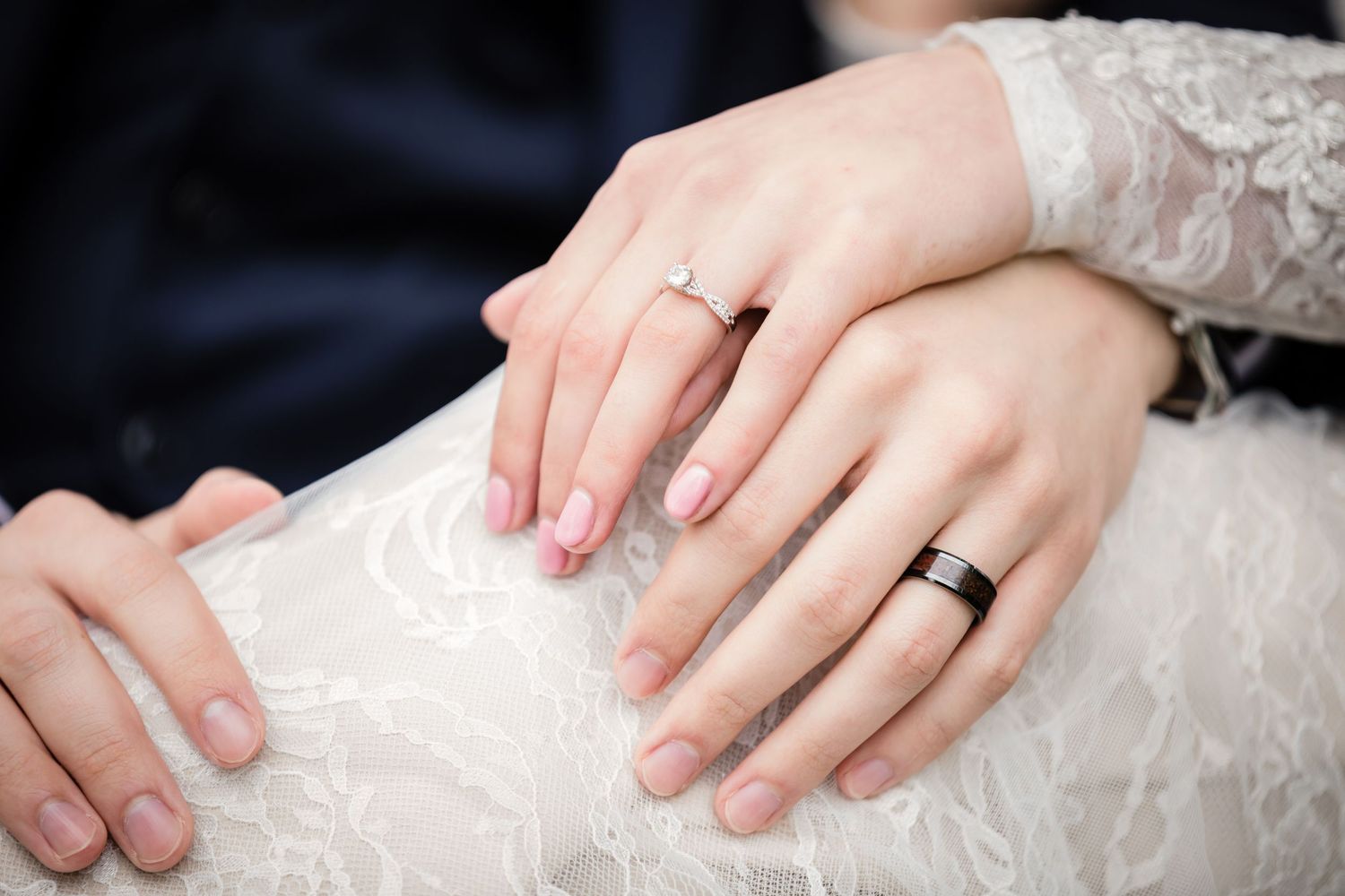 Close-up view of hands wearing wedding rings against white lace fabric showing delicate details and textures.