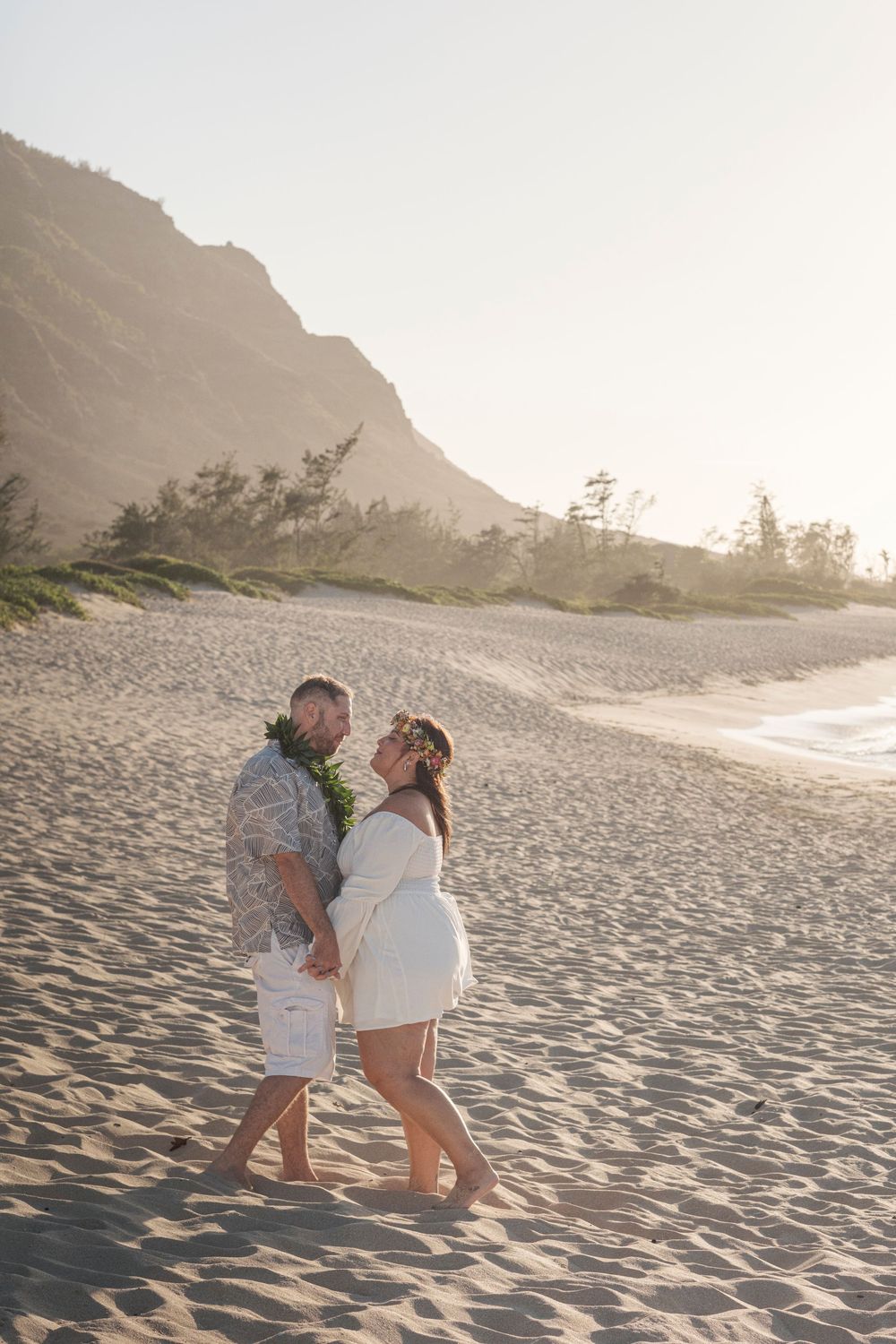 Couple embracing during a North Shore Oahu couples photography session on a sandy beach with mountain views at sunset.