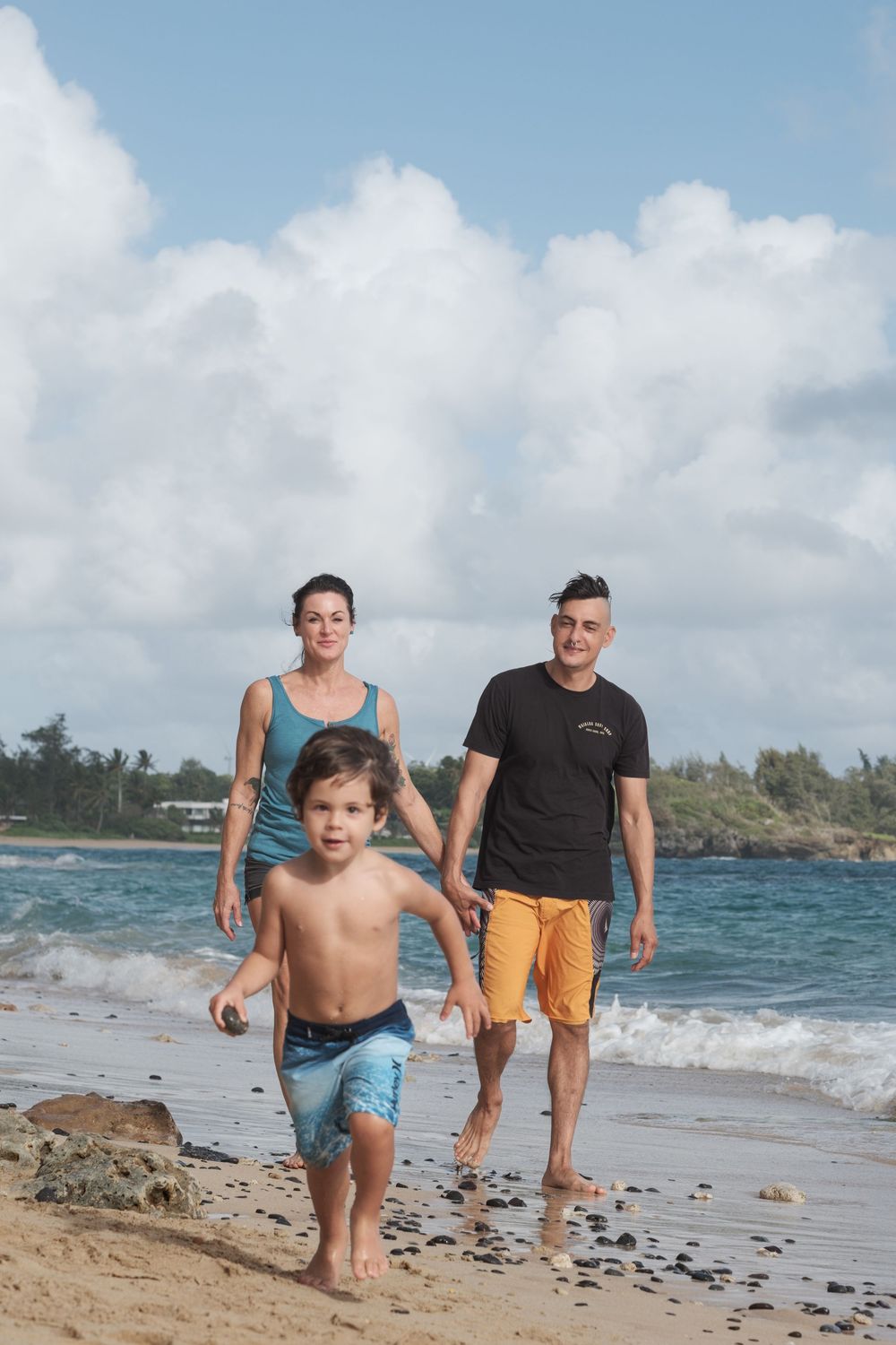 Family walking together along a rocky beach shoreline with turquoise waters and cloudy skies during a family photo session on Oahu.