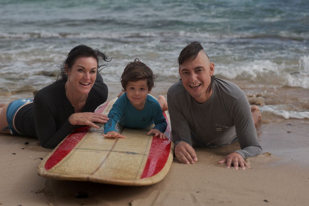 Family of three lying beside a surfboard during a fun North Shore Oahu family photography session by the ocean.
