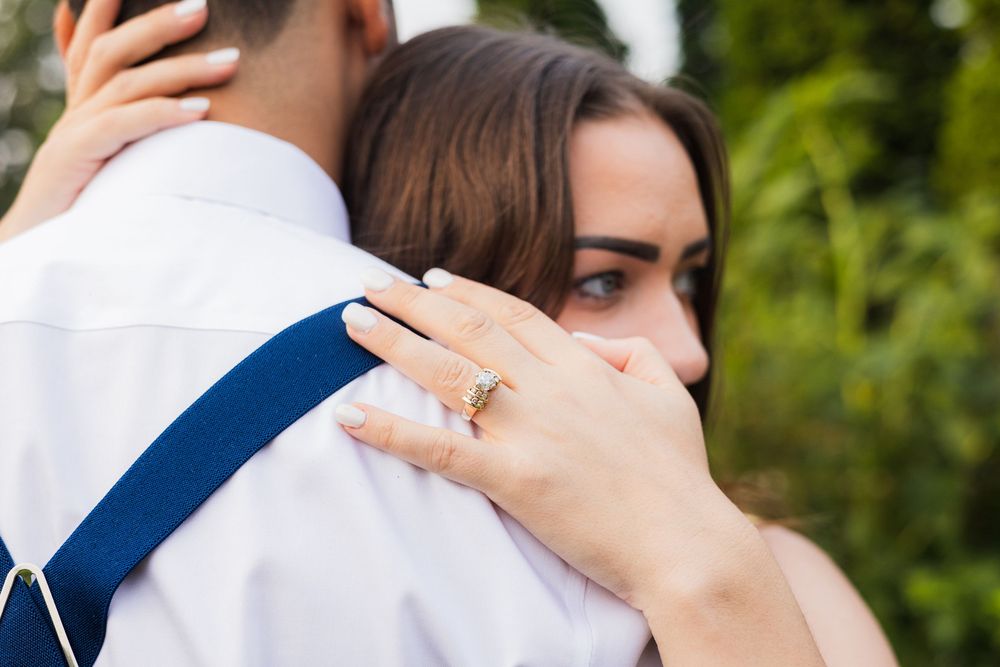 A couple in formal wedding attire embraces while outdoors surrounded by greenery and natural lighting.