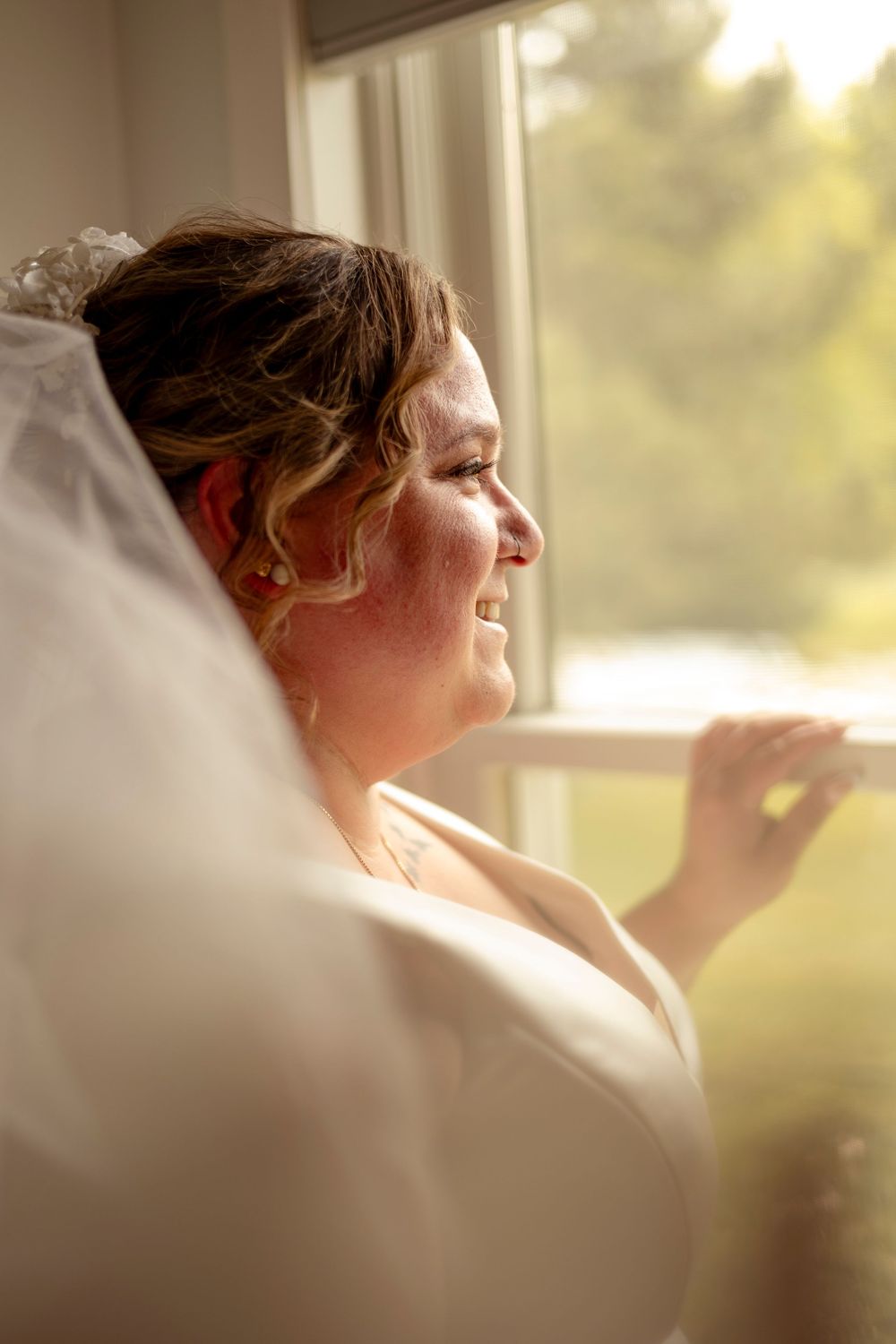 Smiling bride in white dress and veil gazes out a sunlit window on her wedding day.