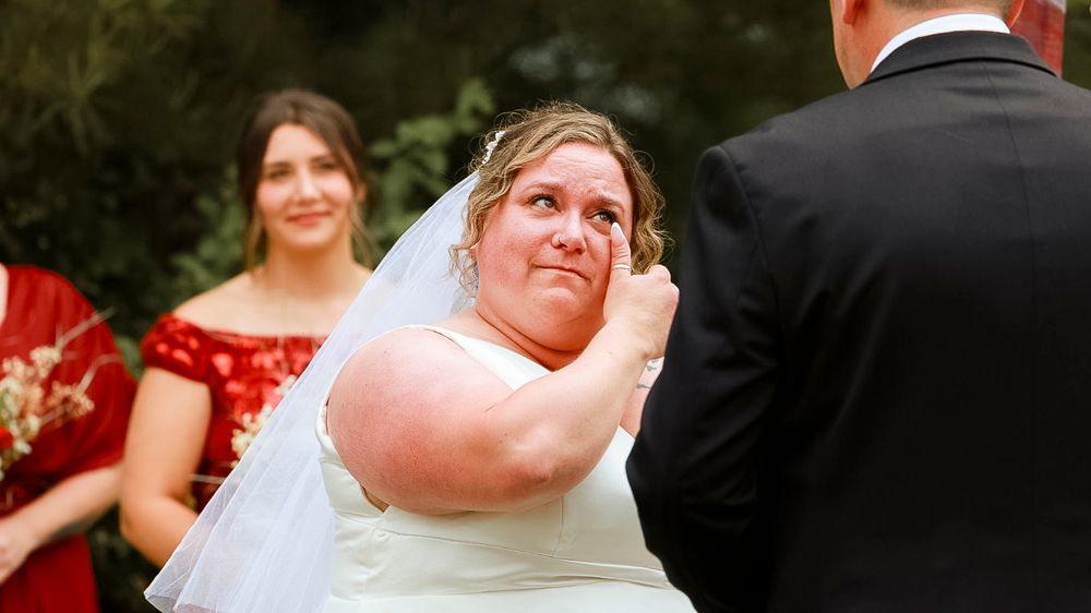 Emotional bride in white gown and veil wiping tears during outdoor wedding ceremony with bridesmaid in red dress behind her.