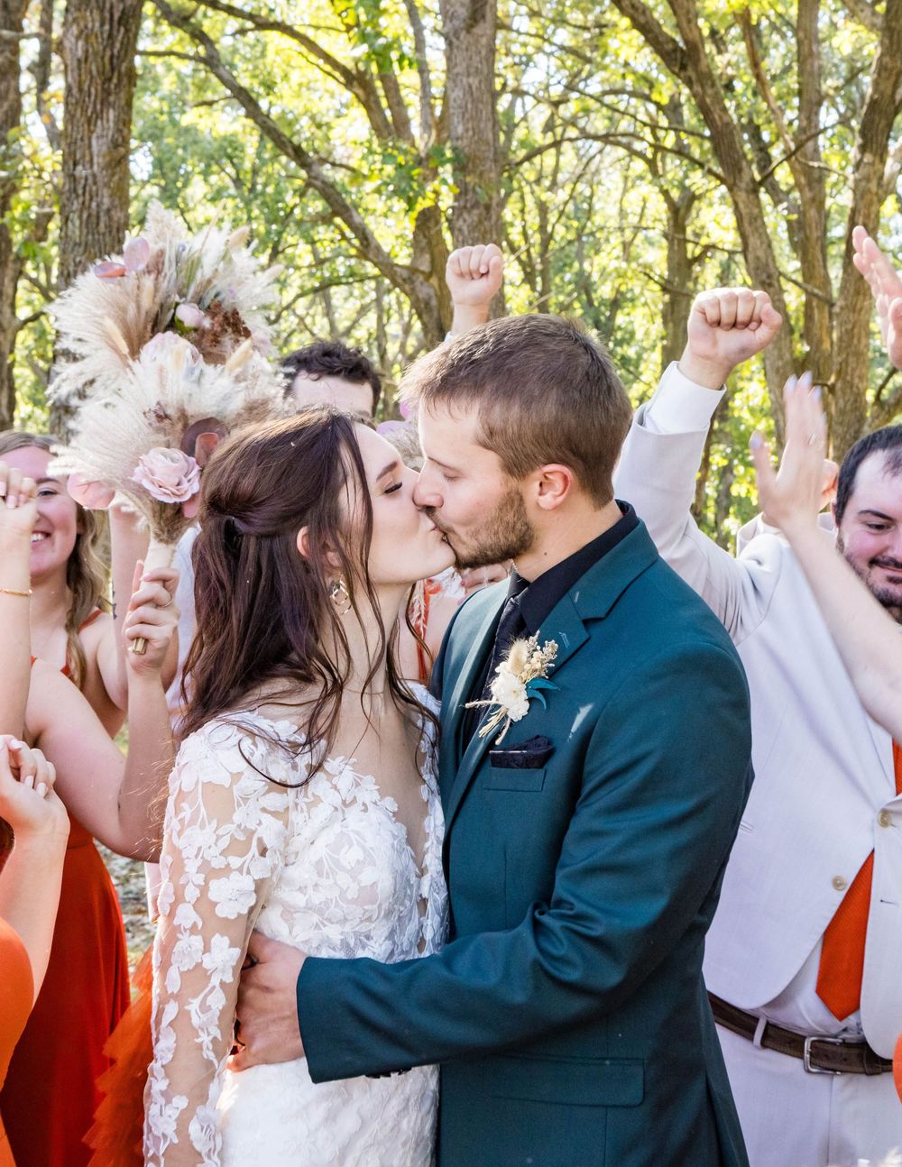 A bride and groom share a kiss surrounded by cheering wedding party members in a lush outdoor forest setting.
