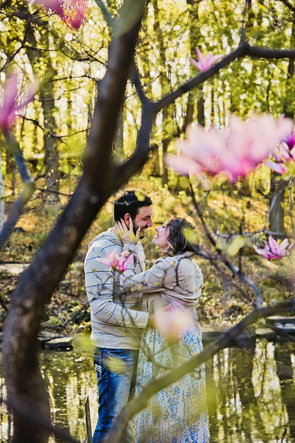Romantic couple embracing near a pond surrounded by blooming pink magnolia trees in a lush spring forest.