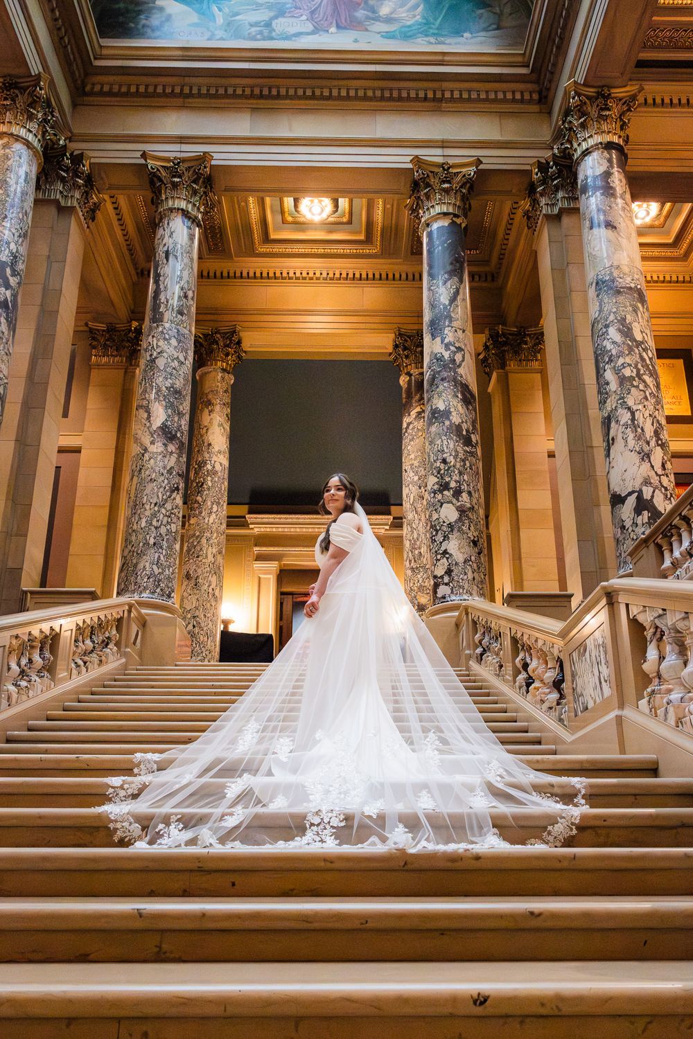 Bride in a flowing white gown with lace veil on grand staircase of ornate building with marble columns.