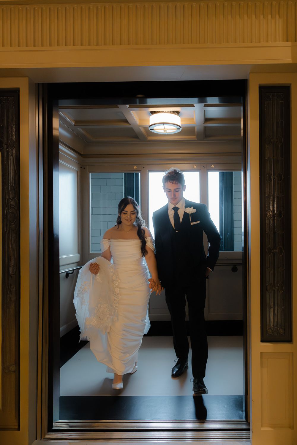 Bride and groom exit elevator holding hands, bride in off-shoulder gown, groom in black suit.