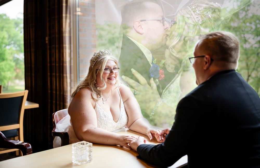 Bride and groom holding hands at a table, with a romantic outdoor kiss reflection in the window behind them.