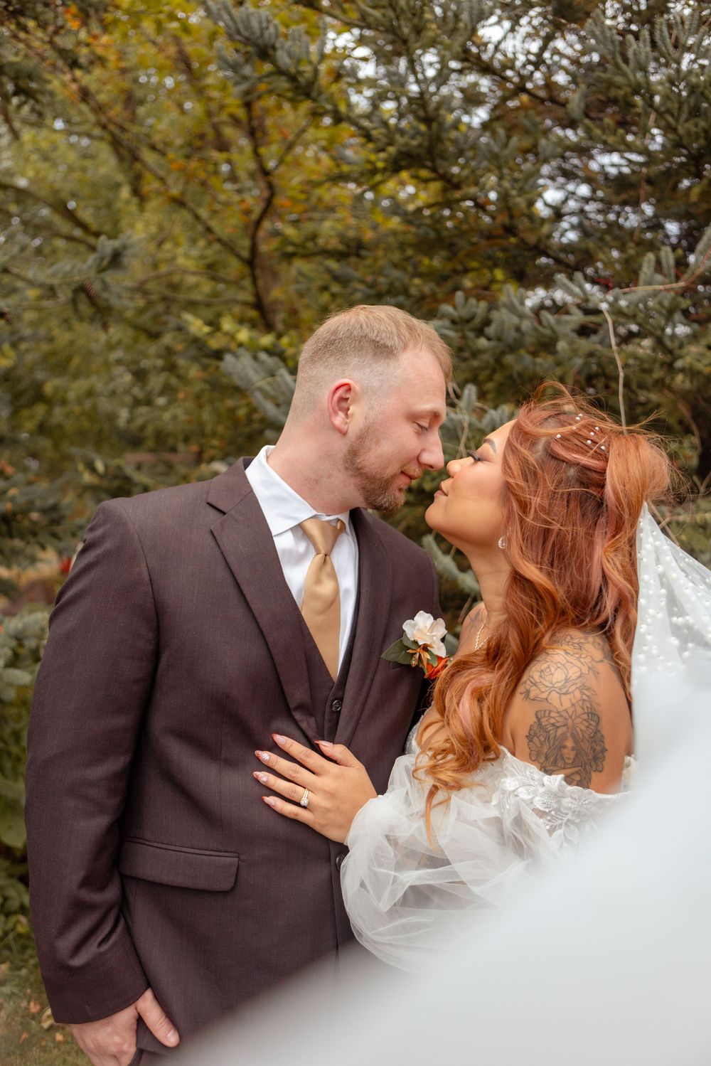 A bride and groom share a romantic moment in an autumn forest, with the bride in a white gown and groom in a brown suit.