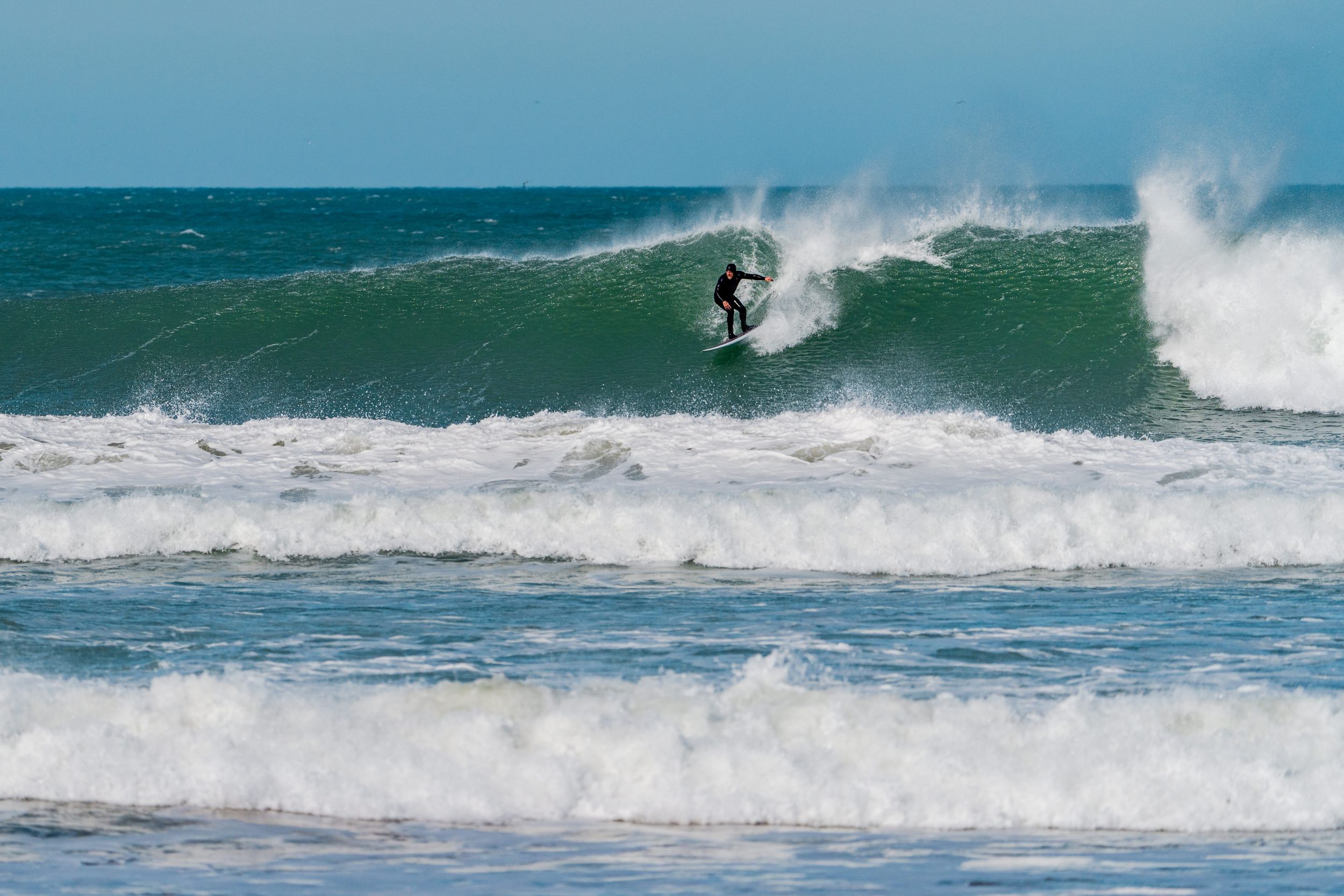 Surfer riding a large turquoise wave with white spray in the ocean on a clear day.
