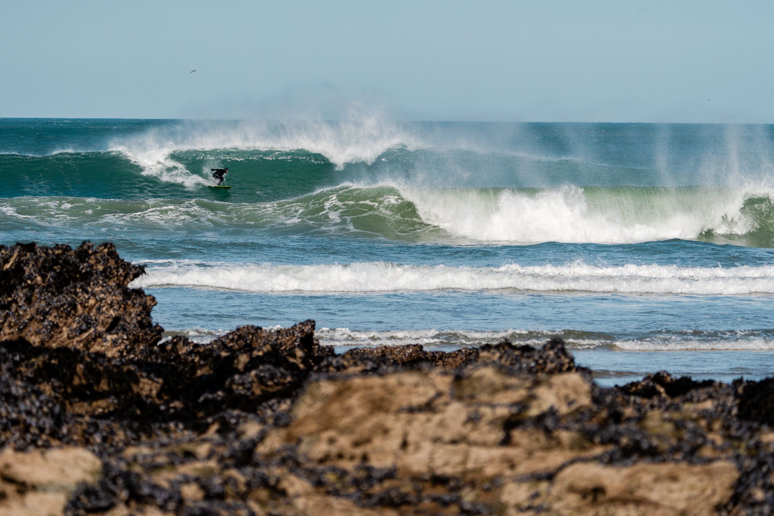 Surfer rides powerful ocean wave with white spray crashing behind on a sunny day at the beach.