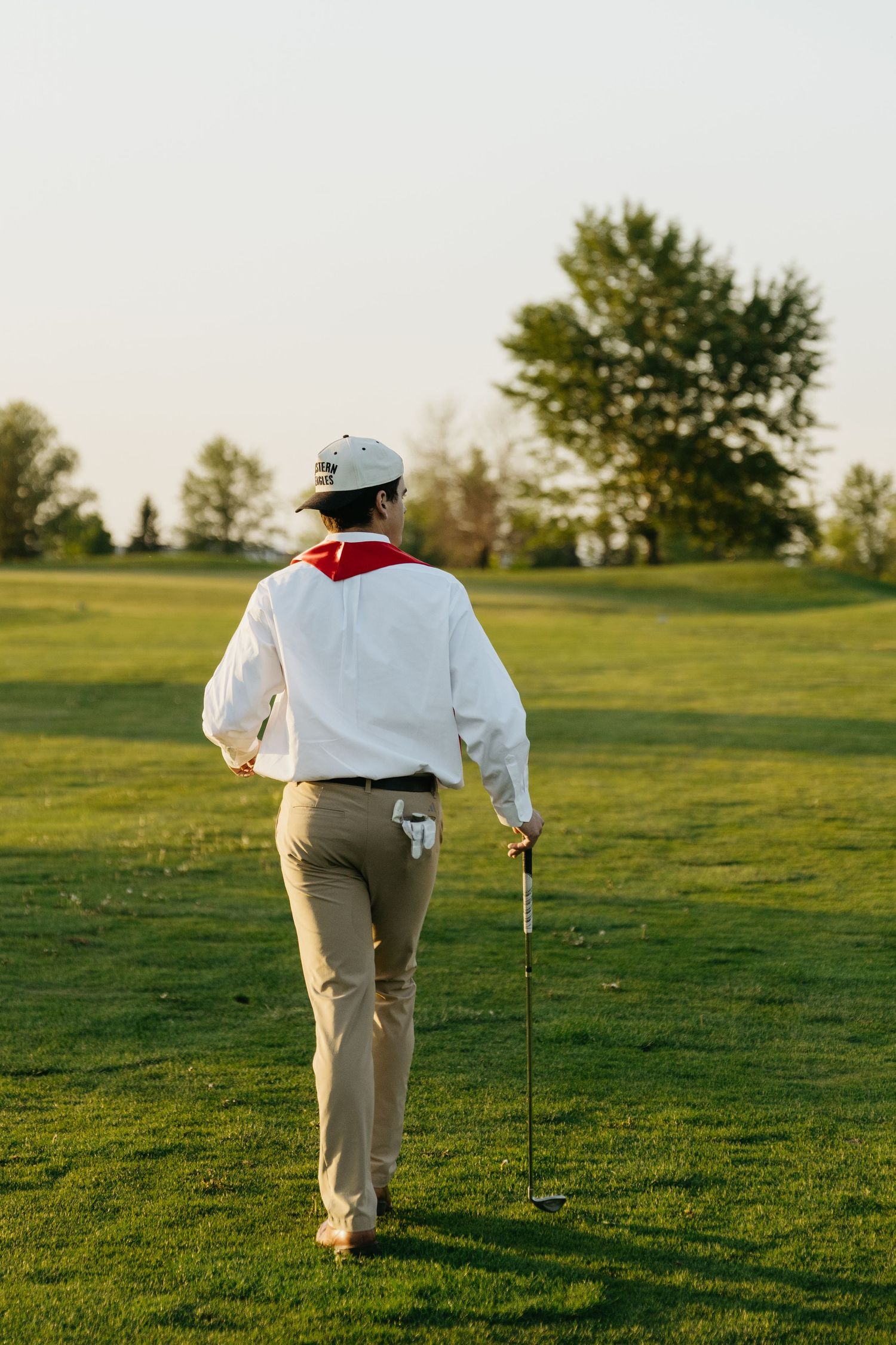 Golfer walking on lush green fairway at sunset, carrying a golf club, wearing white shirt and cap.