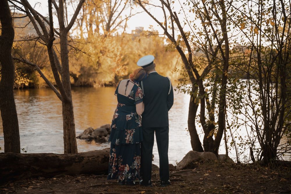 Couple in formal wear embracing by a river surrounded by autumn trees in golden hour light.