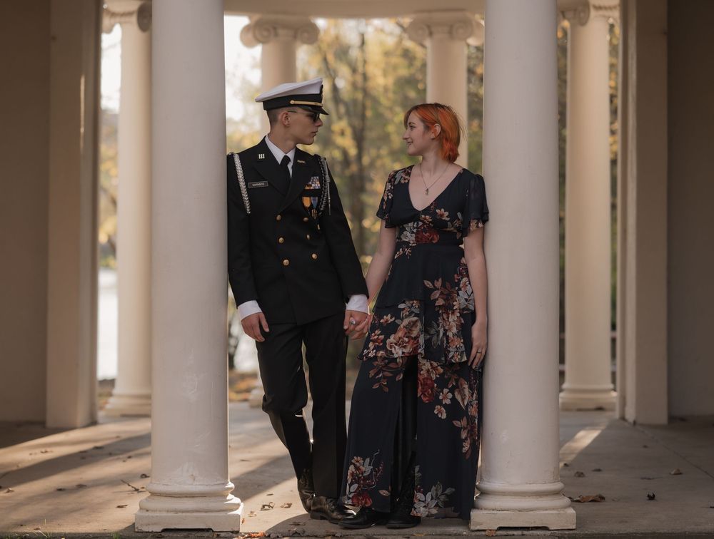 Military officer in dress uniform holds hands with woman in floral gown between white columns outdoors.