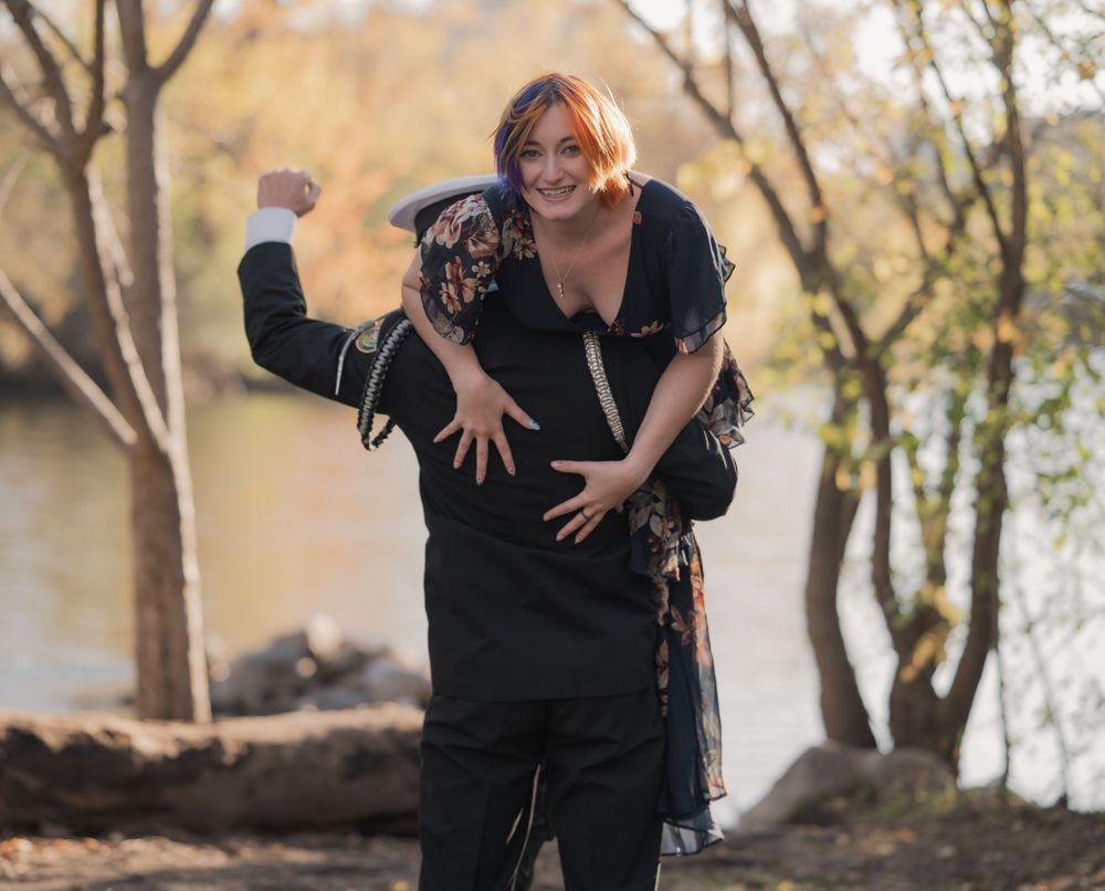 A woman laughs while being carried piggyback by a person in black outdoors near a river with autumn trees.