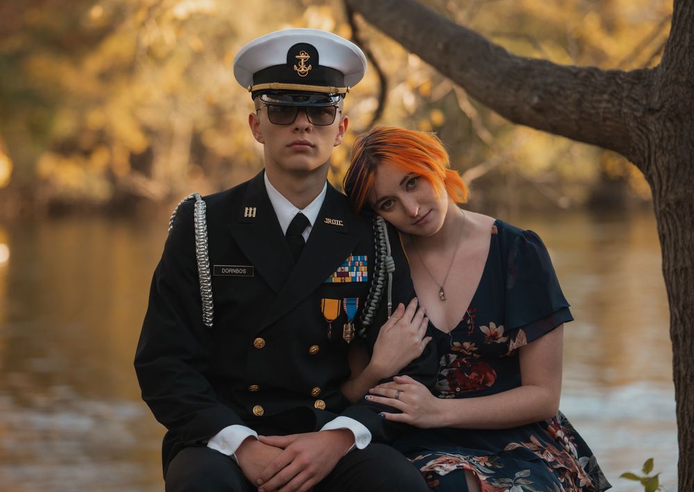 Military couple portrait outdoors, man in Navy dress uniform with medals, woman in floral dress leaning on his shoulder.