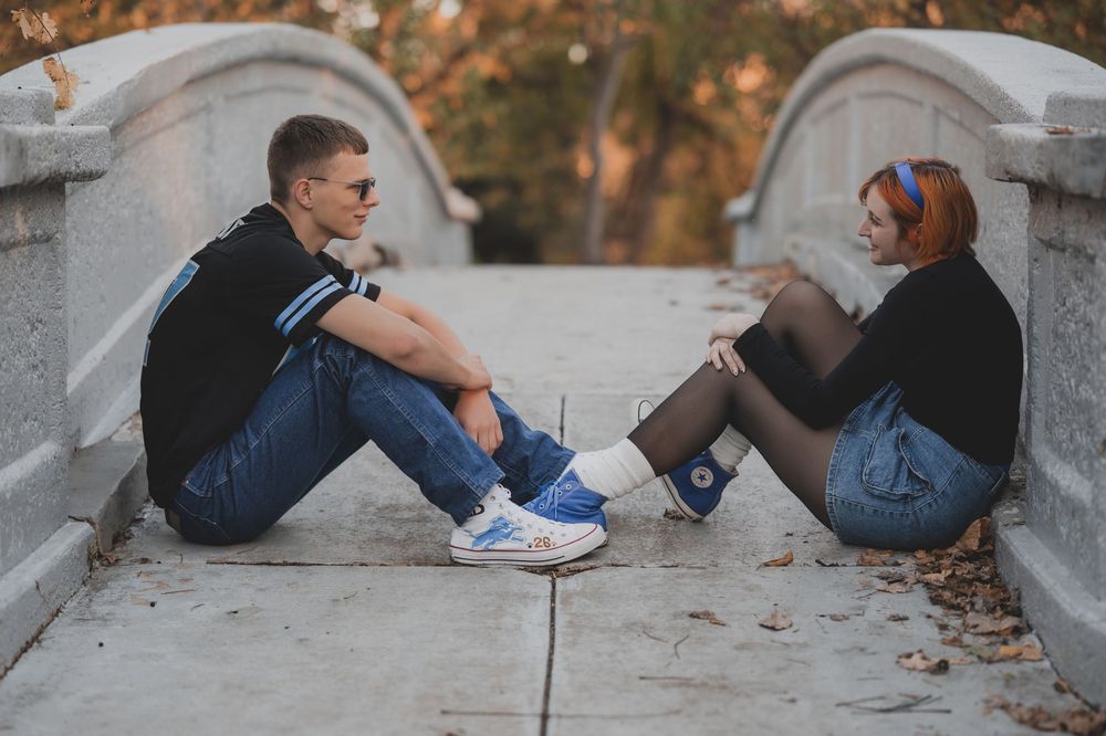 A young couple sits facing each other on a stone bridge pathway in a park during golden hour.
