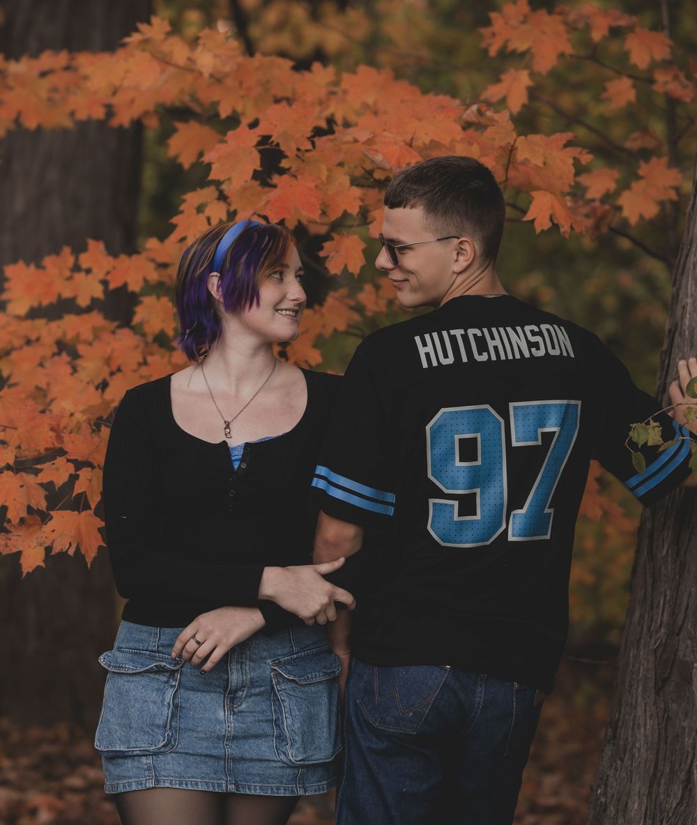 Couple sharing a romantic moment among vibrant orange autumn maple leaves, woman in black top, man in Hutchinson 97 jersey.