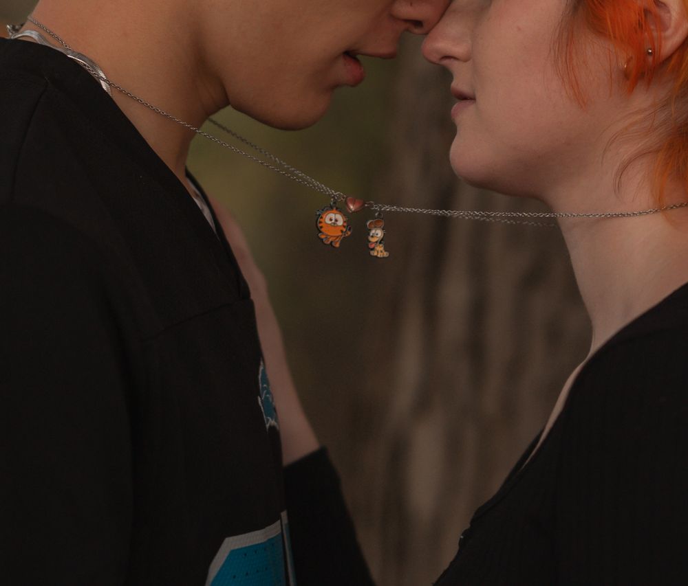 Two teens with necklaces share a romantic moment, faces close together, outdoors with blurred green background.