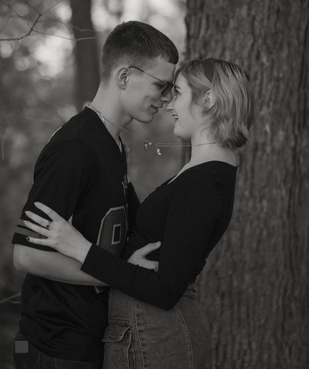 Black and white photo of a young couple embracing and gazing at each other near a tree outdoors.