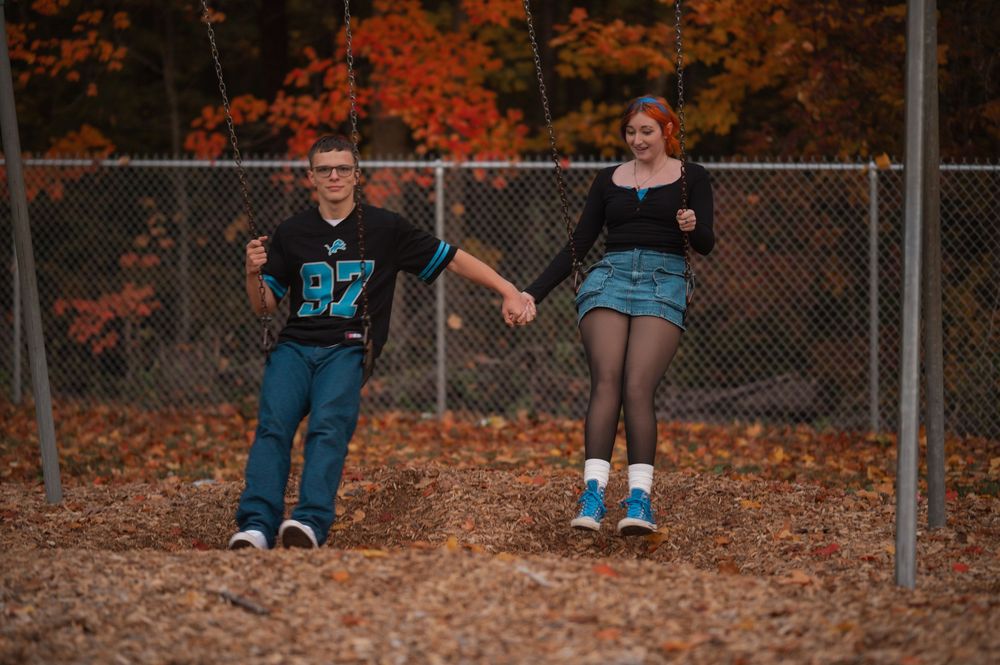 Young couple holding hands in autumn park, wearing casual 90s style outfits near chain-link fence.