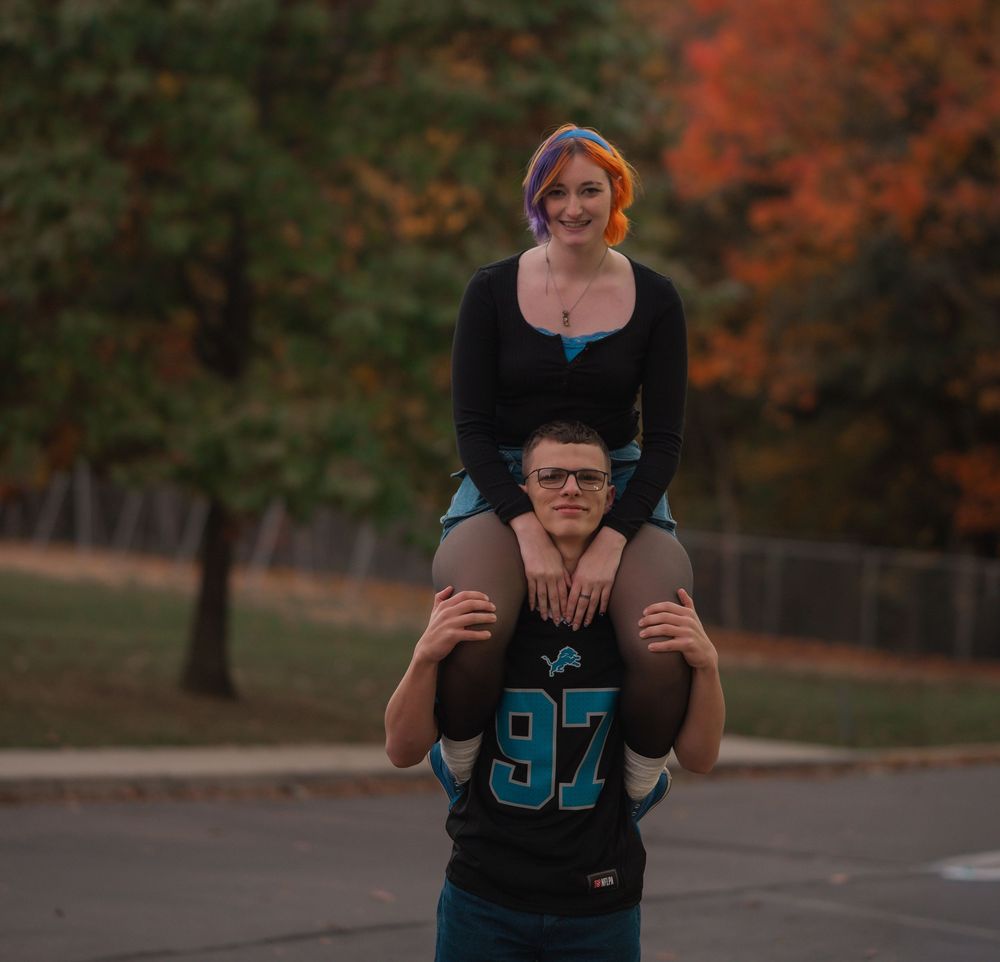 A young woman with colorful hair sits on a man's shoulders outdoors during autumn.