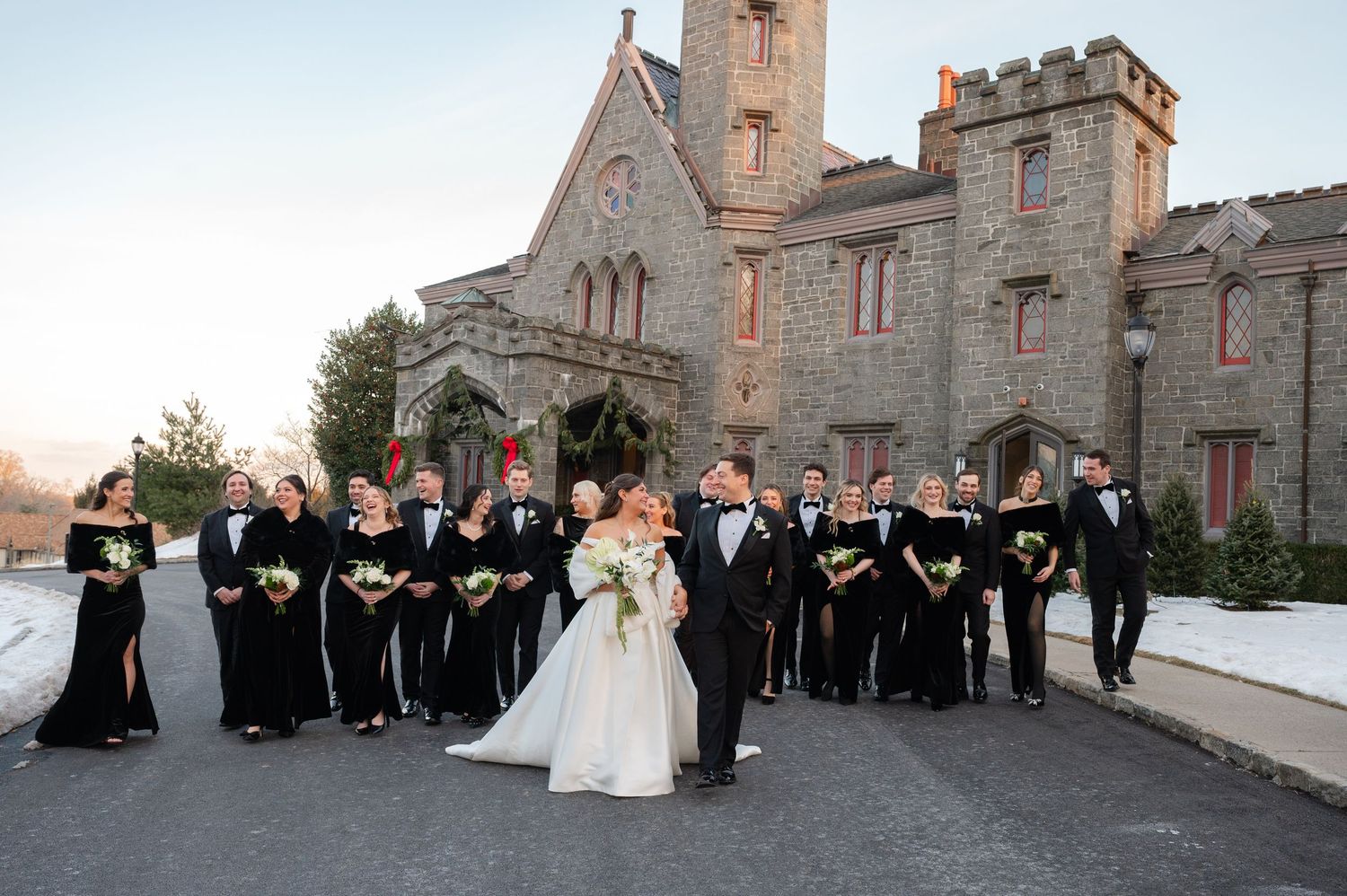 Wedding party in black attire poses outside a stone castle venue, bride and groom sharing a kiss in the foreground.