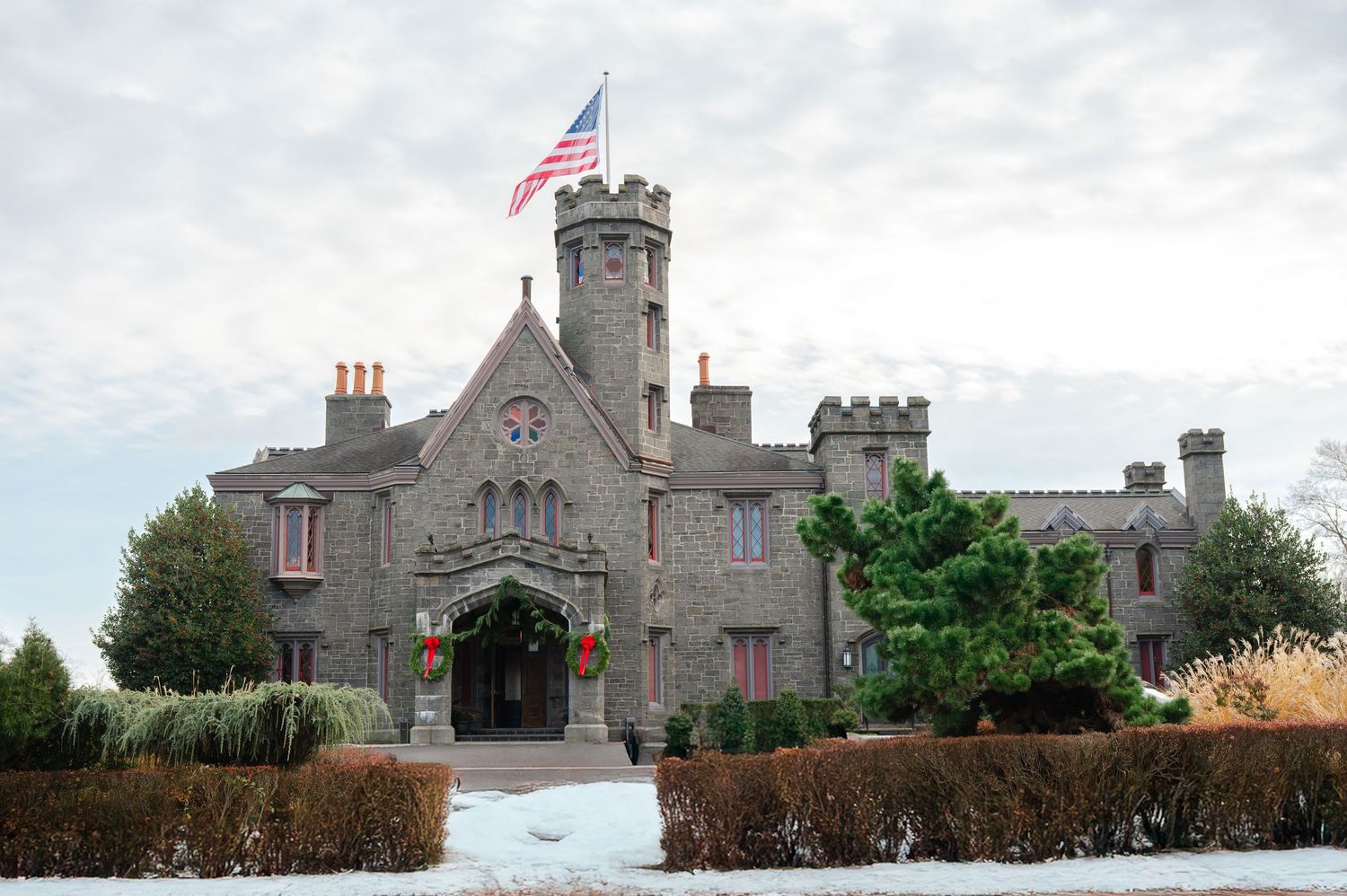 Historic stone castle with American flag, Gothic architecture, snow-covered grounds, and festive holiday decorations at entrance.