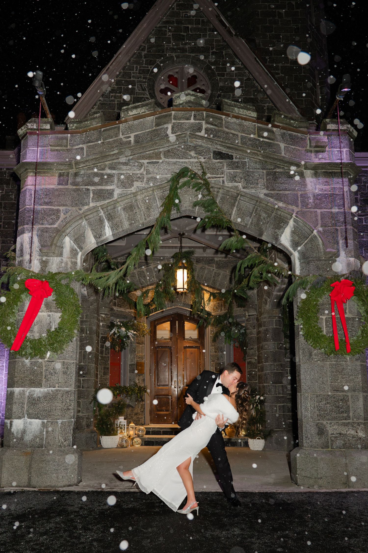 Couple sharing a romantic dip kiss in the snow outside a stone church decorated with Christmas wreaths at night.