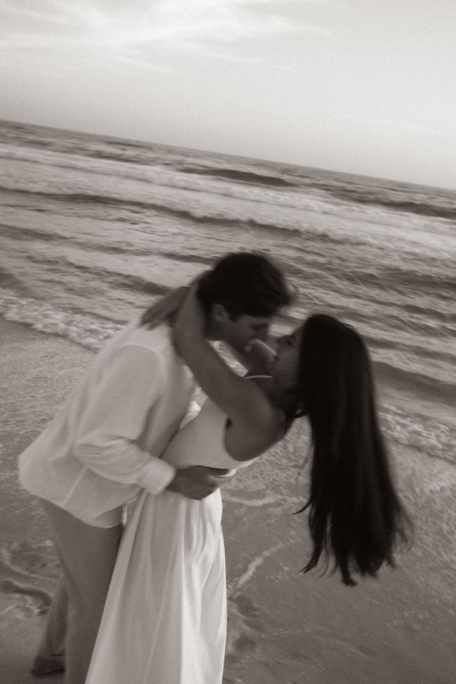 Romantic couple kissing on beach in black and white photo with ocean waves in background.