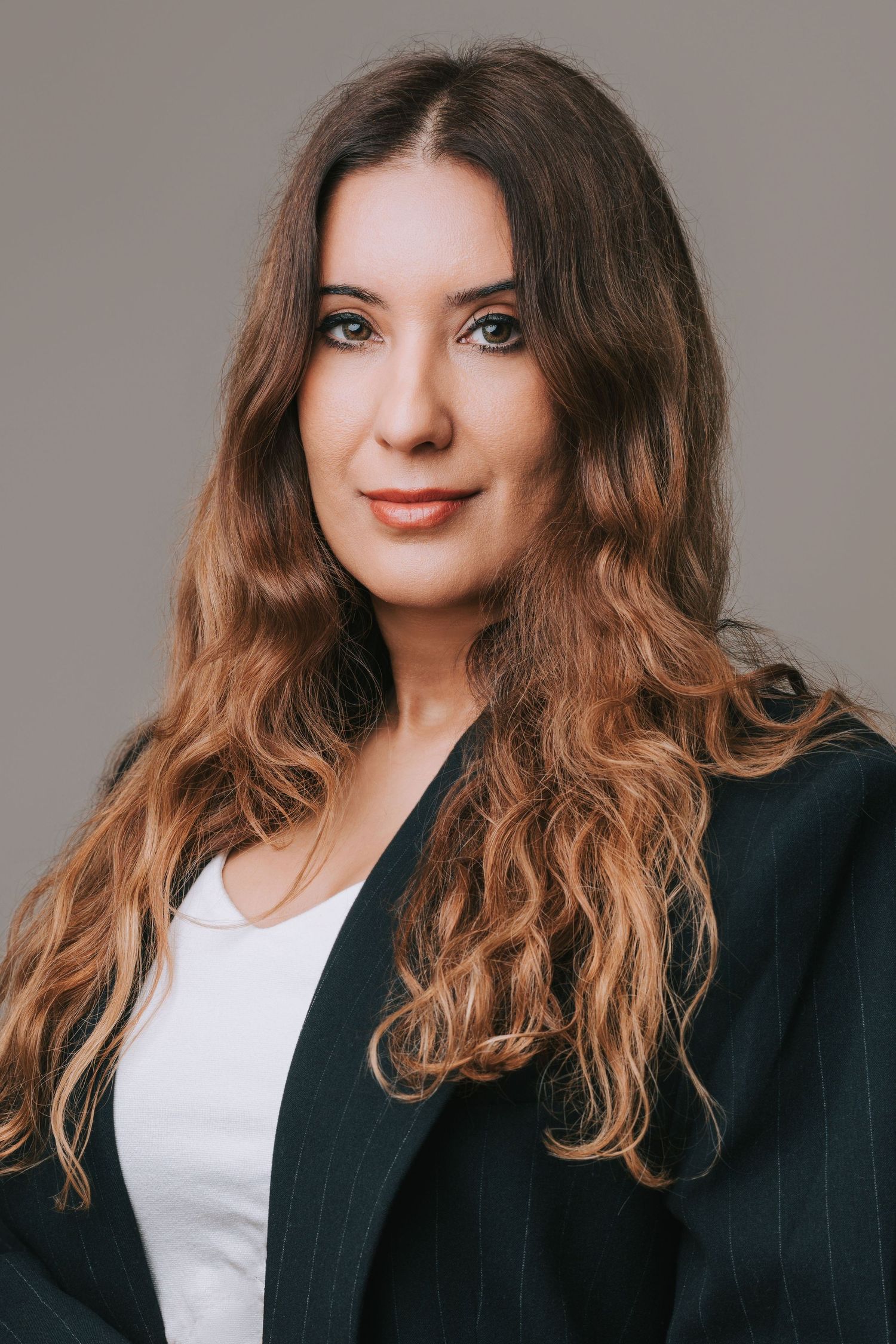 Professional headshot of someone in a dark blazer against black background with soft lighting and wavy brown hair.