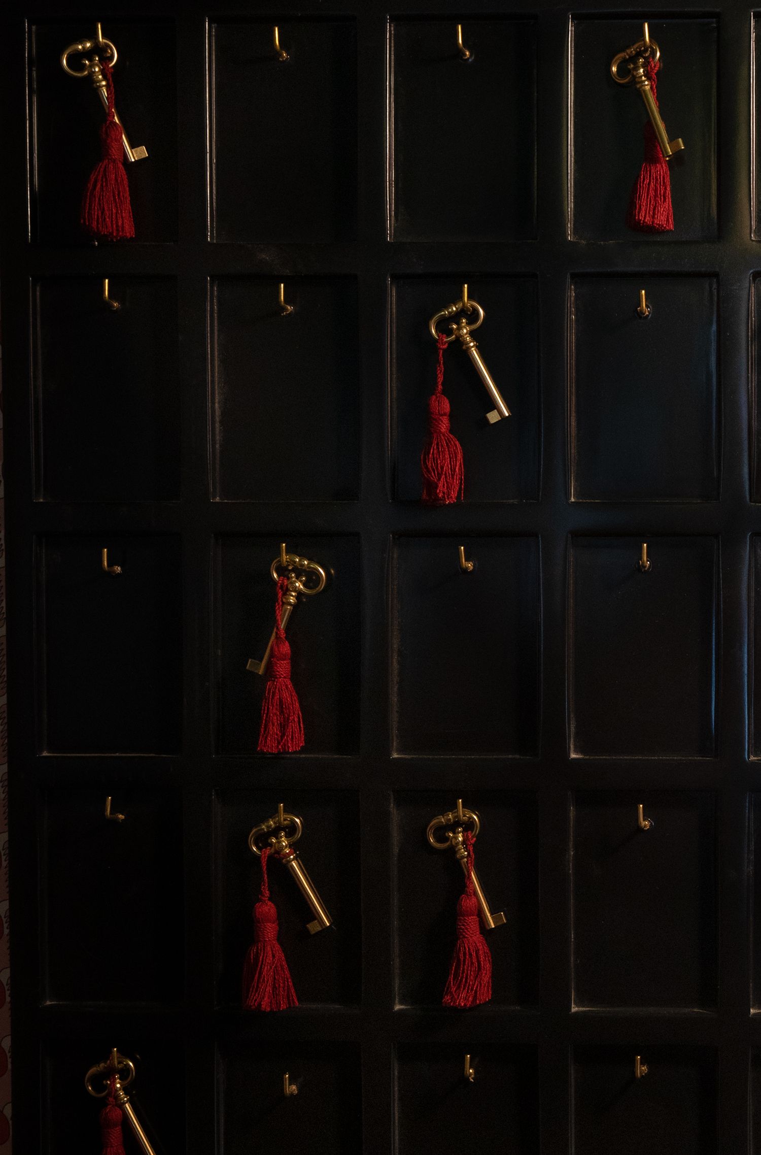 Antique brass keys with red tassels hanging on hooks against a dark wooden display board with grid compartments.