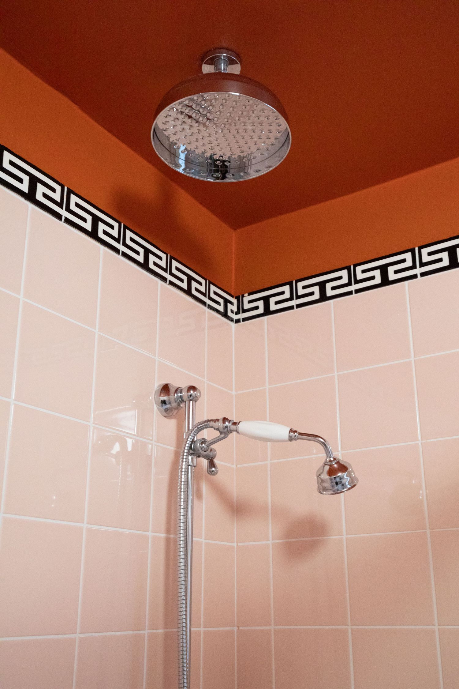 Modern bathroom shower head with white tile walls and black Greek key pattern border trim against an orange ceiling.