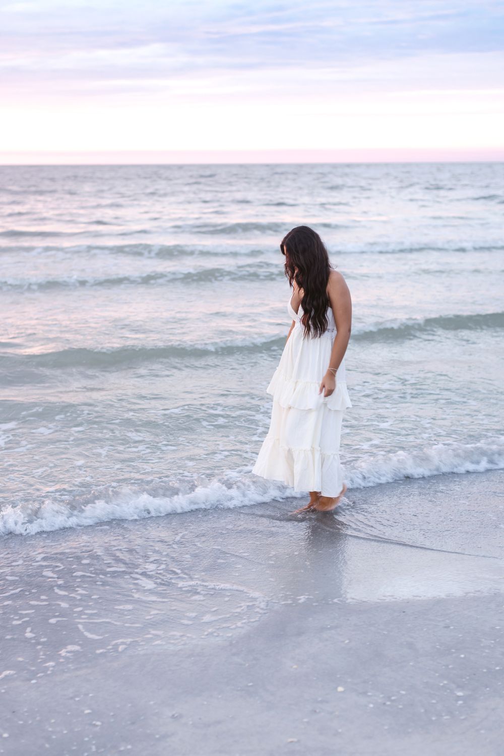 A young lady with long brown hair wears a white dress and walks in calm beach water for senior photos at sunset. Hampton senior photographer