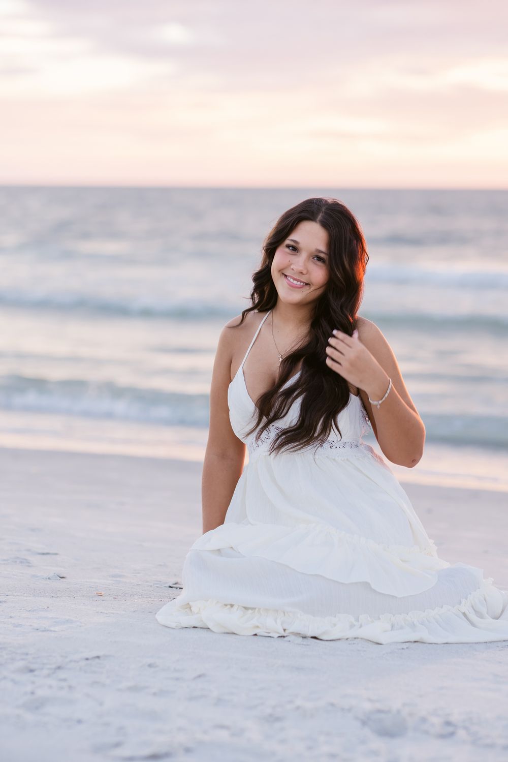 A senior girl wearing white holds her long brown hair from blowing in wind on beach at sunset for senior photos. Kennebunkport senior photos