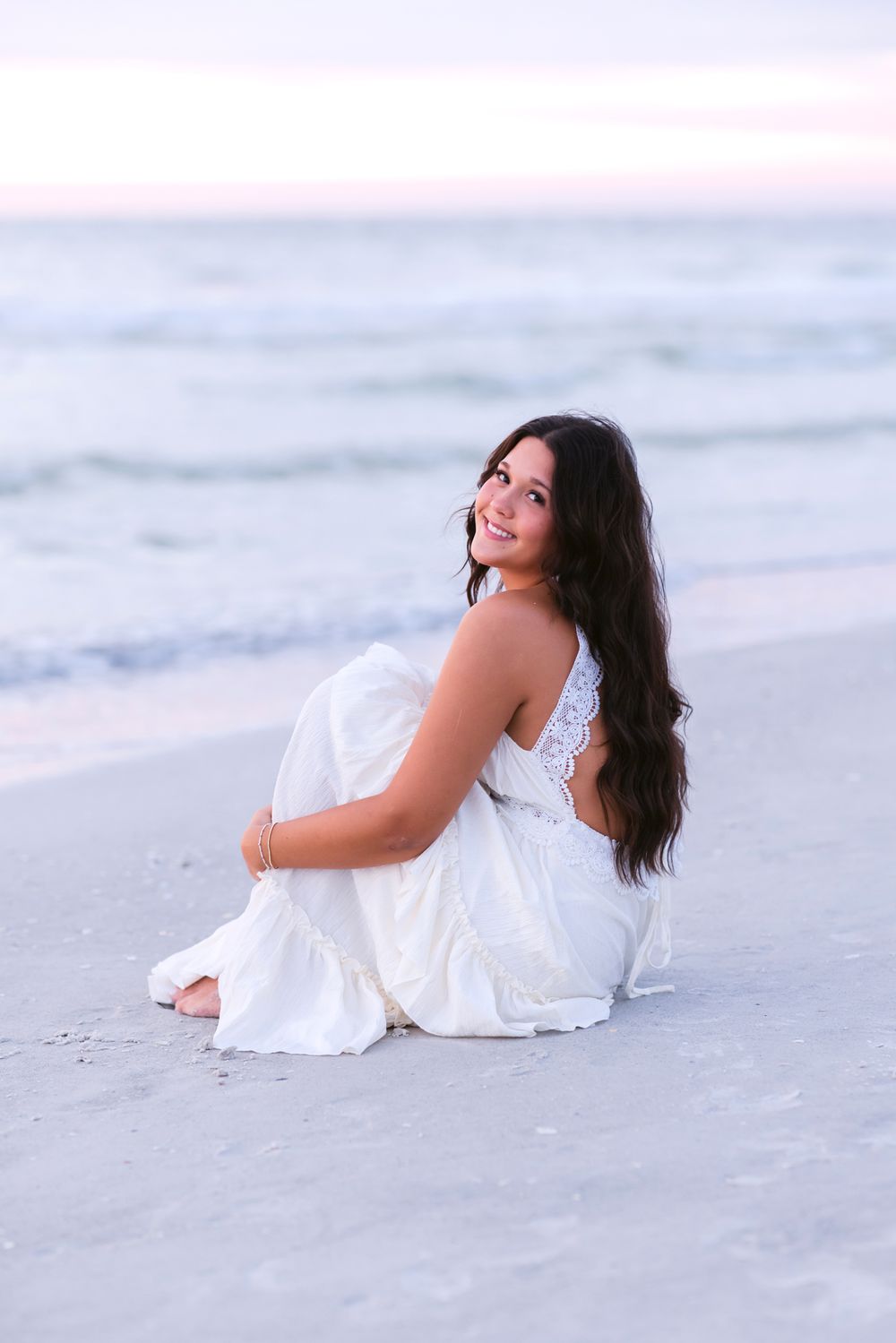 Senior girl wears a white dress and hugs her knees to her chest sitting on Indian Rocks Beach Florida at sunset for senior photos.