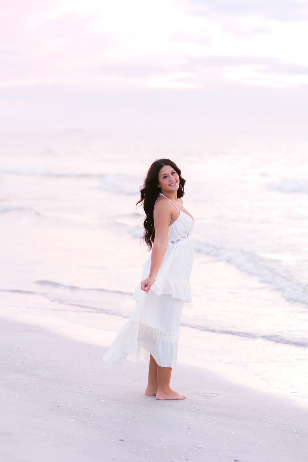 Senior wears a white dress and poses happily on Indian Rocks Beach in Florida at sunset for senior photos. Seacoast NH senior photographer.