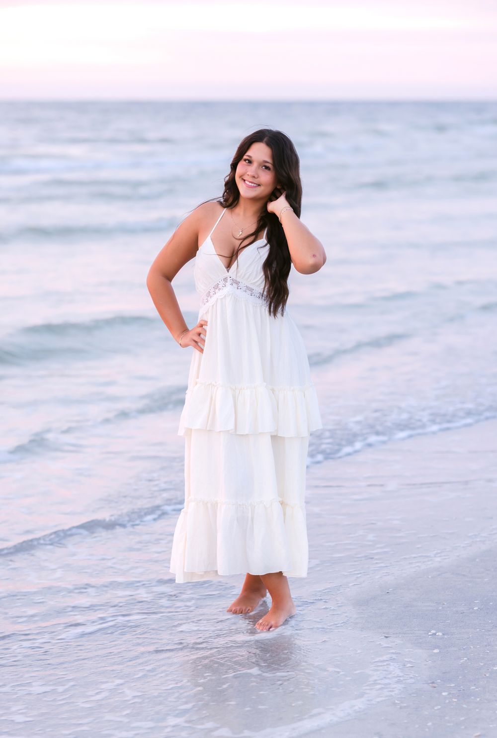 A young lady with long brown hair poses on Indian Rocks Beach at sunset wearing a white dress. North Shore, Cohasset senior photographer.