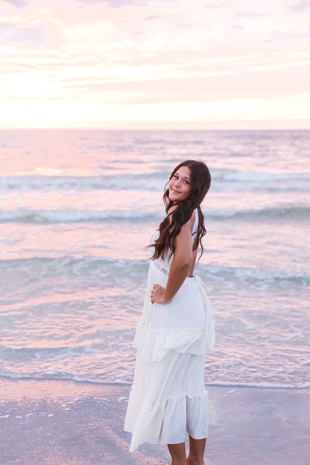 A young lady wears a white dress posing on a beach at sunset at Indian Rocks Beach Florida for senior photos.