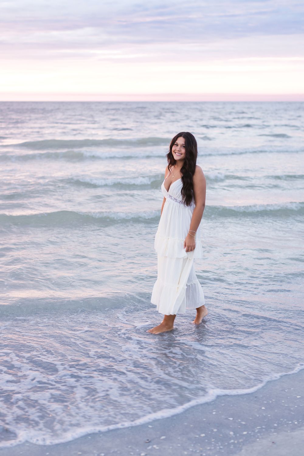 A young lady with long brown hair wears a white maxi dress as she walks through the ocean at sunset for senior photos at Indian Rocks Beach
