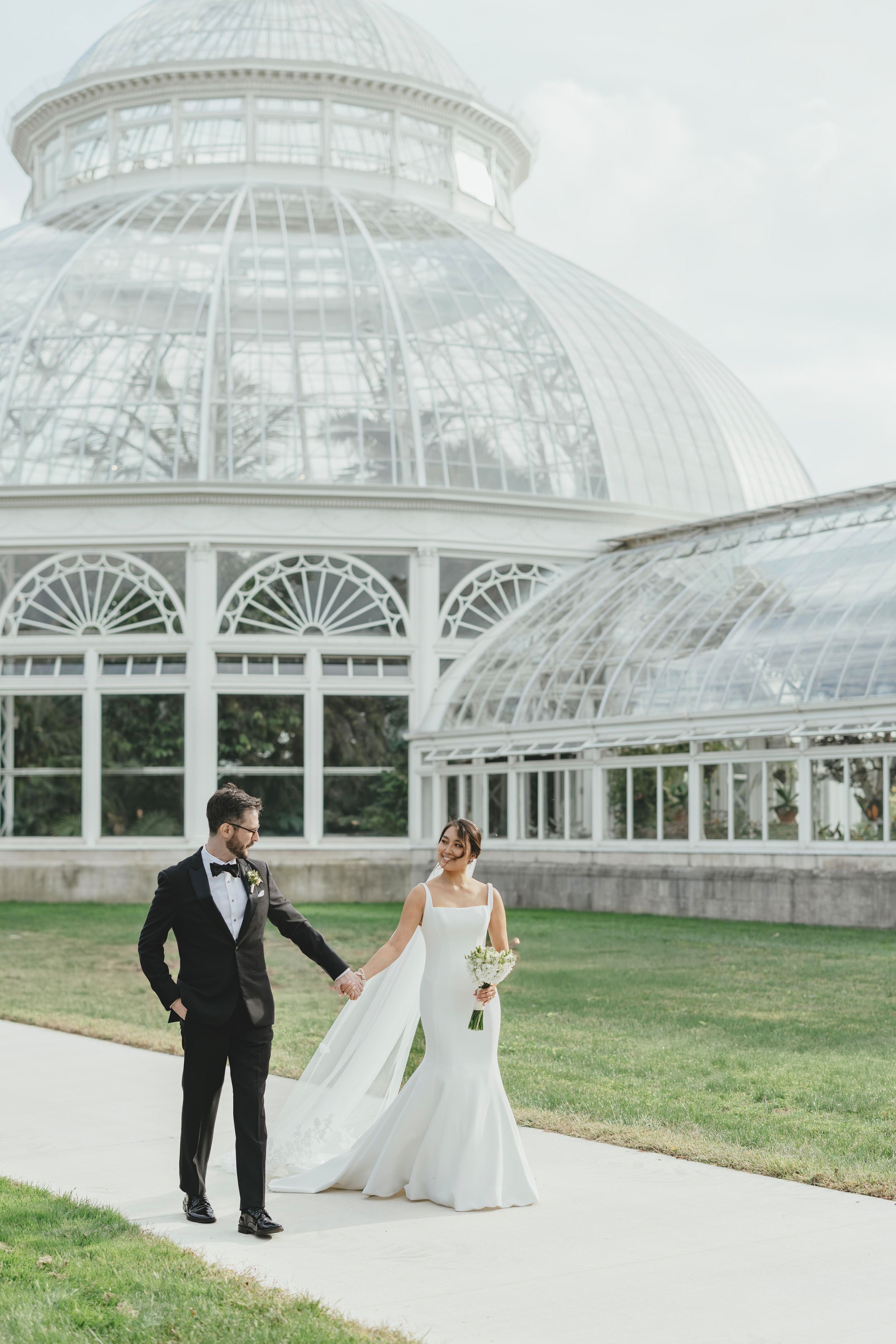 A couple in wedding attire poses for photos outside a grand glass conservatory building with ornate architecture.