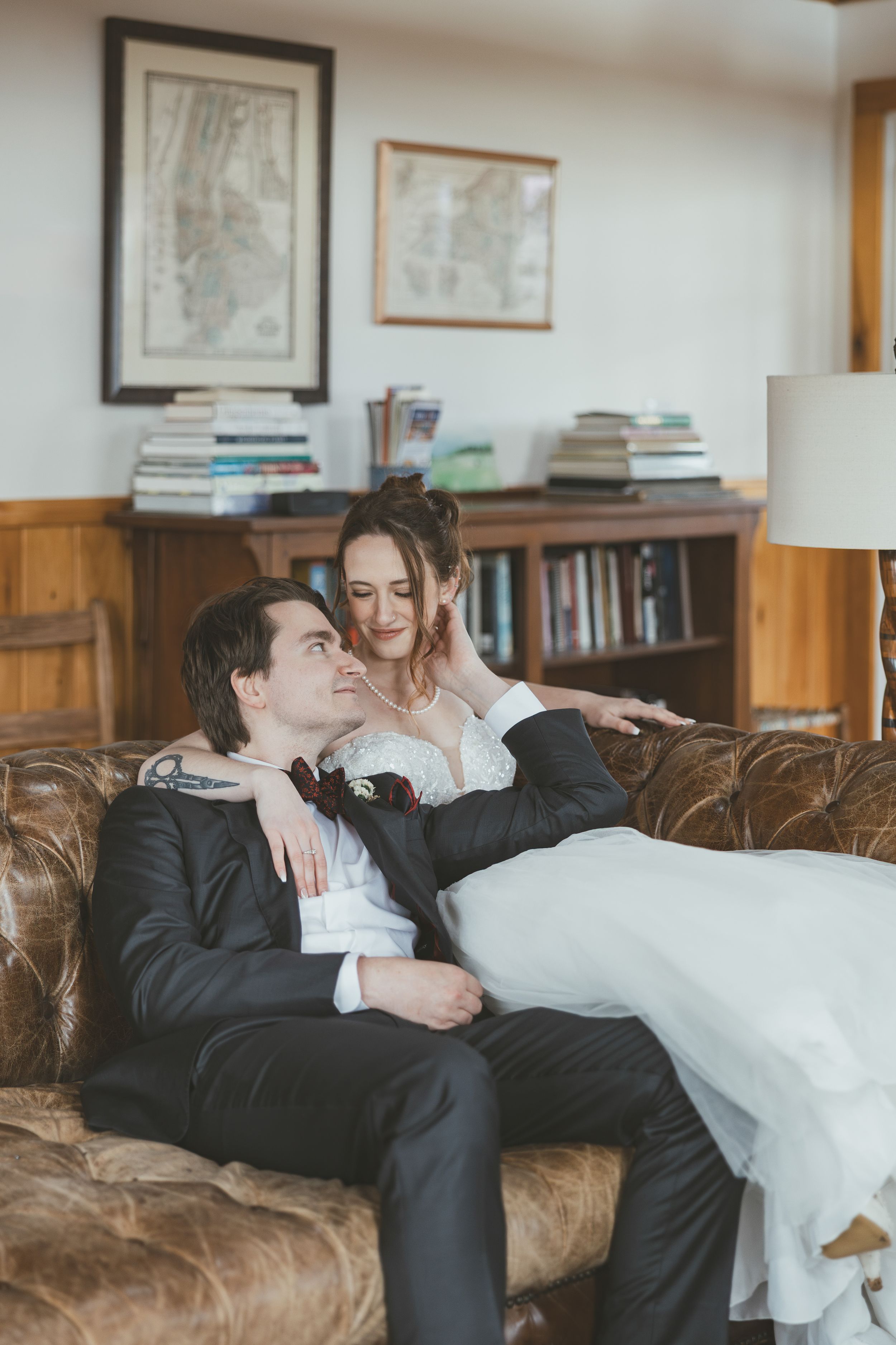A couple in wedding attire shares tender moments while relaxing together on a brown leather couch.