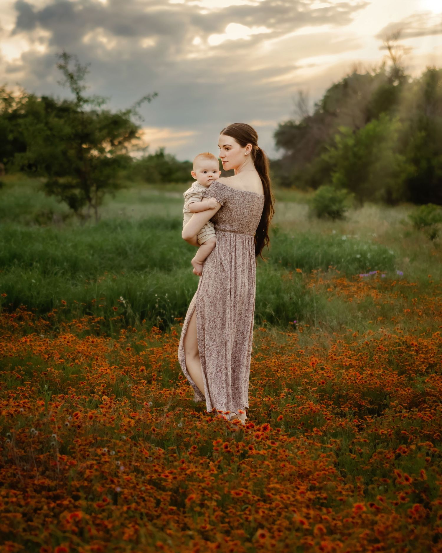 A woman in a flowing maxi dress stands in a field of vibrant orange wildflowers at sunset while holding her baby close in Fort Worth, TX