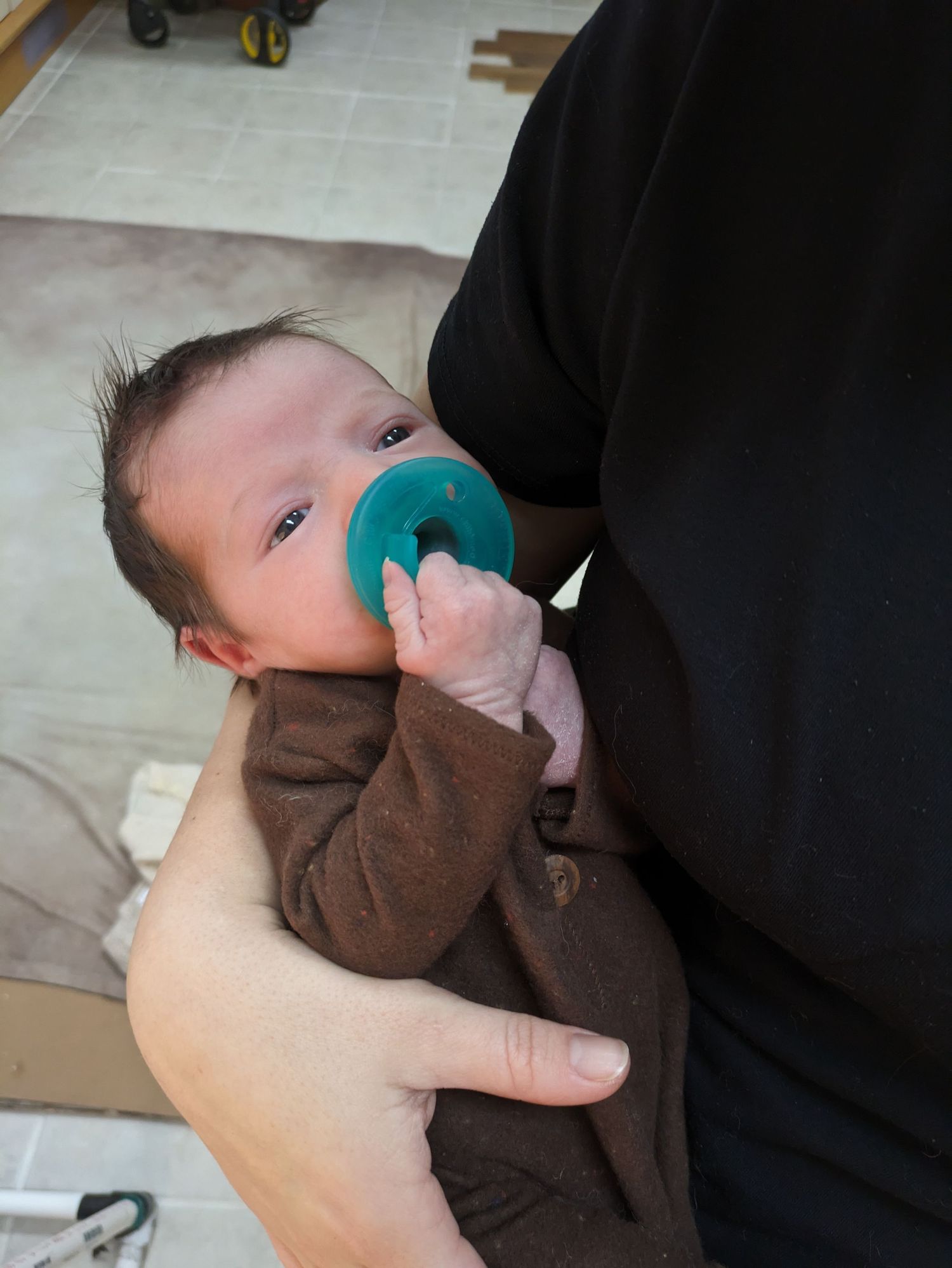 A baby holds a teal-colored teething toy while being cradled in brown fabric.