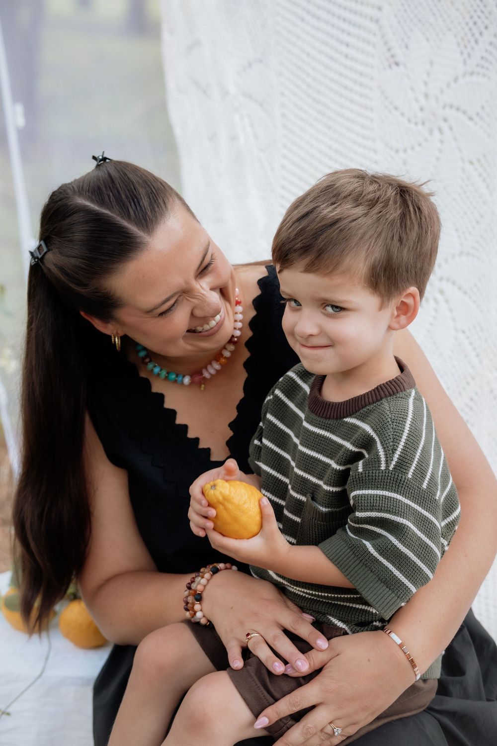 A tender moment shared between parent and child as they sit together with a citrus fruit against a white lace backdrop.