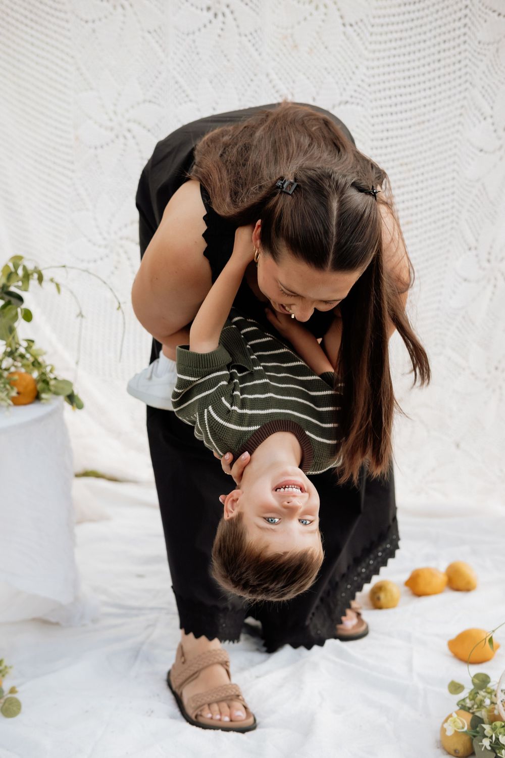 Playful upside-down moment captured between parent and child with oranges scattered on white fabric.