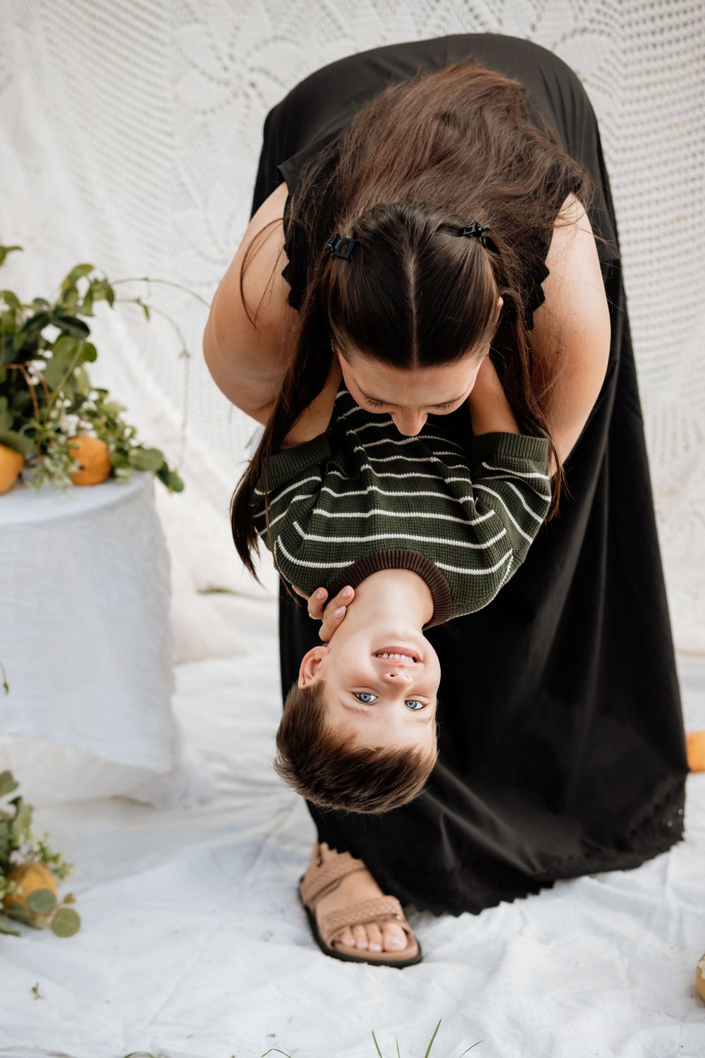 Playful upside-down moment between parent and child against white backdrop with greenery.